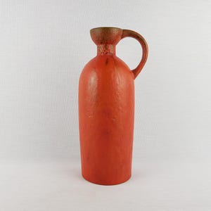 Puede incluir: Una jarra alta de cerámica naranja con asa. La jarra tiene un cuerpo redondeado y un cuello estrecho que se ensancha en la parte superior. El asa y la parte superior de la jarra son de color marrón oscuro. La jarra probablemente se usa para contener líquidos.