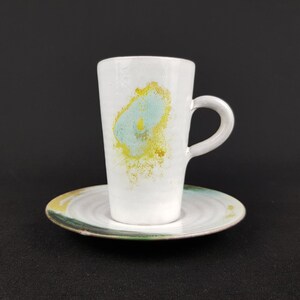Peut inclure: Un ensemble tasse et soucoupe en céramique blanche composé d'une tasse haute et conique avec une anse incurvée. La tasse présente une éclaboussure abstraite bleu clair et jaune. La soucoupe assortie a une texture striée avec des touches vertes.