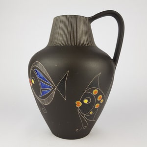 May include: A black ceramic pitcher with a handle, featuring two stylized fish designs. One fish has blue and white details, the other has orange and yellow accents. The top has vertical lines.