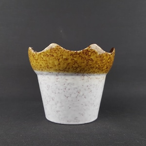 Op de afbeelding: Een kleine keramische bloempot met een golvende, getextureerde rand. Het bovenste gedeelte is bruin en geel gespikkeld, terwijl de body wit is met bruine spikkels. De pot heeft een licht uitlopende vorm.
