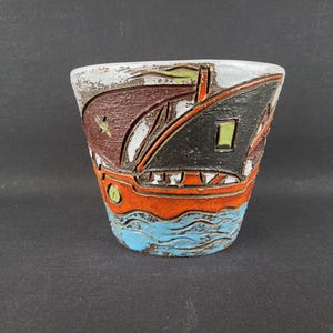Könnte beinhalten: Ein Keramikübertopf mit einem strukturierten, handbemalten Design eines Bootes auf dem Wasser. Das Gefäß zeigt einen braunen und schwarzen Rumpf, einen leuchtend orangefarbenen Mittelteil und hellblaue Wellenmuster an der Basis mit einem grünen Akzent.