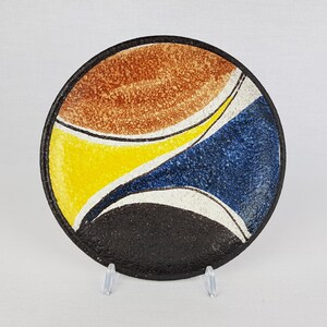 May include: A round ceramic plate with a black, brown, yellow, and blue abstract design. The plate has a textured surface.