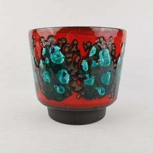 Puede incluir: Una maceta de cerámica con un exterior rojo vibrante, adornada con detalles turquesa y marrón. La maceta tiene forma cilíndrica con una base negra. Adecuada para plantas de interior.