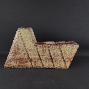 Könnte beinhalten: Eine Keramikvase mit einer kantigen, asymmetrischen Silhouette und einer strukturierten, gesprenkelten Oberfläche in Creme- und Rotbrauntönen. Die Oberfläche ist durch markante, vertiefte diagonale Linien gekennzeichnet, die ein segmentiertes, geometrisches Erscheinungsbild erzeugen.