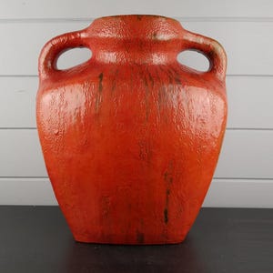 Puede incluir: Un jarrón grande de cerámica naranja con dos asas en forma de bucle. El jarrón tiene una superficie texturizada y un acabado brillante. El color es un naranja rojizo vibrante con vetas más oscuras, lo que le da un aspecto rústico. Es un objeto decorativo.