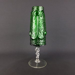 Puede incluir: Un jarrón de vidrio verde con una superficie texturizada y un tallo de vidrio transparente. El jarrón tiene un diseño único con círculos en relieve y un tallo retorcido.