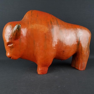 Op de afbeelding: Handgemaakte, oranje gekleurde bizonskulptuur. De sculptuur heeft een getextureerd oppervlak en staat in een staande positie. De bizon heeft een gedetailleerde kop en lichaam, met zichtbare hoorns. Het kunstwerk is een decoratief stuk.