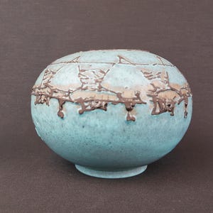 Könnte beinhalten: Eine runde, türkisfarbene Keramikvase mit strukturierter Oberfläche und einem dunkelbraunen Zierband in der Mitte. Die Vase steht auf einem kleinen, kreisförmigen Sockel. Das Design ist organisch und künstlerisch.