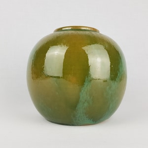 Puede incluir: Un jarrón redondo de cerámica verde con un acabado brillante. El jarrón tiene un efecto degradado sutil, pasando de un verde más oscuro en la parte inferior a un verde más claro en la parte superior.