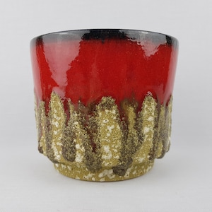 Op de afbeelding: Een keramische plantenbak met een glanzend rode bovenkant en een getextureerde, aardse basis. De basis heeft een patroon van verticale, ruw bewerkte ribbels in tinten bruin, beige en wit. De plantenbak heeft een zwarte rand.