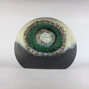 May include: A semi-circular ceramic vase with a textured surface. The base is a solid matte black, transitioning upwards into a cream-coloured top. A prominent circular pattern features concentric rings of dark brown, vibrant green, and white.