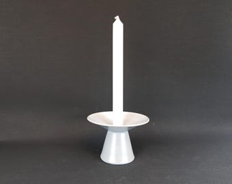 May include: A white candle sits on a silver candlestick. The candlestick has a wide, flat base and a narrow, cylindrical stem.