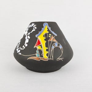 Könnte beinhalten: Eine schwarze Keramikvase mit einem farbenfrohen, abstrakten Hausdesign. Das Haus hat gelbe, rote und blaue Akzente. Ein weißes Blumenmuster verziert die Seite der Vase. Die Vase hat eine abgerundete Form und steht auf einer weißen Oberfläche.