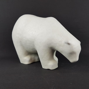 Può includere: Scultura in ceramica bianca di un orso polare in posizione accovacciata. L'orso guarda in basso e sembra annusare il terreno.