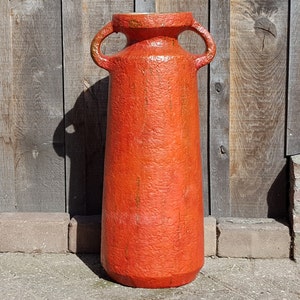 Puede incluir: Un jarrón alto de cerámica naranja con dos asas. El jarrón tiene una superficie texturizada y una base ligeramente acampanada.