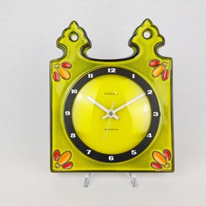 May include: A square wall clock with a lime green ceramic frame featuring decorative orange and red floral motifs in the corners. The clock face is bright yellow with a black circular border, white numerals, and the text Kienzle electronic.