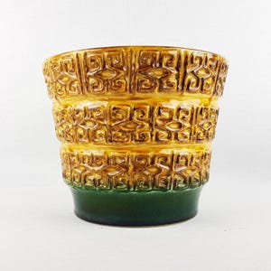 May include: A ceramic planter featuring a tiered, cylindrical shape with a glossy finish. The upper two-thirds display a raised, repeating geometric pattern in shades of golden yellow and brown, while the base is a solid, smooth dark green.