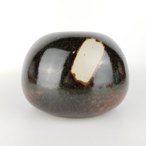 May include: A rounded, ceramic vase with a glossy, dark brown finish. A lighter, rectangular patch of off-white color is visible on the top. The base transitions to a reddish-brown hue.