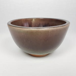 Puede incluir: Un cuenco de cerámica marrón con una abertura ancha y una base redondeada. El cuenco tiene un acabado liso y brillante con un degradado de colores, que va del marrón oscuro a un marrón verdoso más claro cerca de la base. El cuenco tiene unos 15 cm de diámetro.