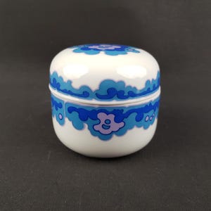 May include: A small, white ceramic jar with a lid. The jar and lid are decorated with a blue and purple floral design. The jar is round and has a smooth, glossy finish. The background is a solid black.