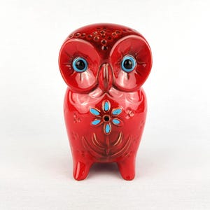 Puede incluir: Figura de búho de cerámica roja brillante, con ojos azules y un diseño floral turquesa en el pecho. El búho tiene una superficie texturizada y detallada y se apoya en tres pequeñas patas. Artículo decorativo.
