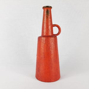 Puede incluir: Un jarrón de cerámica rojo con una superficie texturizada y un asa. El jarrón tiene un cuello estrecho y una base ancha.