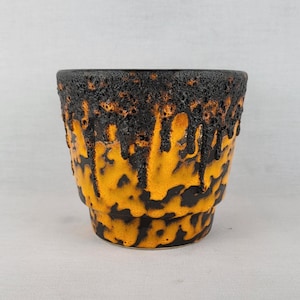 May include: A ceramic planter featuring a vibrant orange base color with a thick, textured black glaze dripping from the rim. The surface has a porous, lava-like appearance at the top, transitioning into a smooth, mottled orange and black pattern.