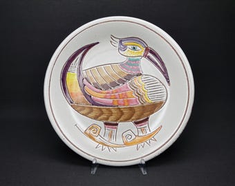 May include: A white ceramic plate with a colorful stylized bird design. The bird is brown, yellow, orange, pink, and purple with a green eye. The bird is perched on a branch with stylized leaves.