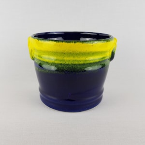 Puede incluir: Una maceta de cerámica con base azul oscuro, que transiciona a un borde amarillo y verde. La maceta tiene un acabado brillante y una forma simple y redondeada. Adecuada para plantas pequeñas o suculentas.