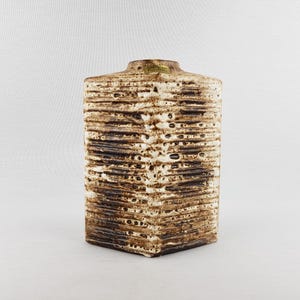 May include: A rectangular ceramic vase with a textured, layered design in shades of brown and cream. The vase has a square base and a slightly flared top. The surface has a rough, organic feel, with dark brown accents.