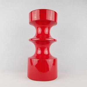 Puede incluir: Un jarrón de cerámica de color rojo brillante con un acabado lustroso. El jarrón presenta una silueta escultural distintiva con una base cilíndrica ancha, una sección central estrecha y un borde superior acampanado, creando un aspecto escalonado.