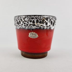 Op de afbeelding: Een keramische plantenbak met een felrode body en een getextureerde zwart-witte rand. De plantenbak heeft een bruine basis en een kleine, ovale sticker met de tekst "Topeko Keramik".
