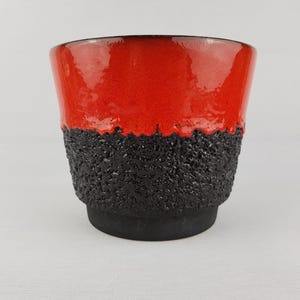 May include: A ceramic planter featuring a vibrant, glossy red upper section that transitions into a textured, matte black base. The surface of the lower portion has a rough, granular appearance, while the top rim is smooth and reflective.