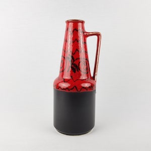 May include: A tall, ceramic vase with a handle. The top portion is red with a black, patterned design, while the base is black. The vase has a sleek, elongated shape, suitable for home decor.