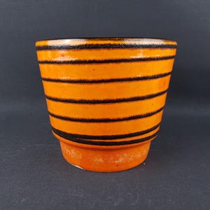 Puede incluir: Maceta de cerámica naranja con rayas horizontales negras. La maceta tiene forma cilíndrica con una base ligeramente ensanchada. El interior y la base son de un tono naranja más claro.