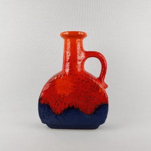 May include: A ceramic vase with a handle, featuring a vibrant color scheme. The top and handle are a bright orange, transitioning to a deep red, and finishing with a dark blue base. This decorative piece is suitable for home decor.