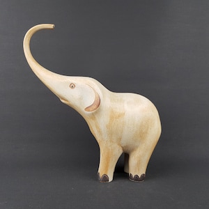 May include: A cream-colored elephant figurine with a smooth, polished finish and subtle tan shading. The elephant is depicted in a standing position with its trunk raised high, featuring dark brown accents on the feet and inner ear details.
