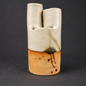May include: A handmade ceramic vase with a unique design. The upper portion is a light beige color with two cylindrical openings. The lower section is a warm orange-brown. Dark brown drips and speckles add character to the piece.