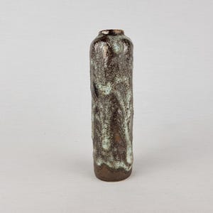 May include: A tall, cylindrical ceramic vase with a narrow opening. The surface features a dark brown base color with a textured, mottled glaze in shades of light blue and white that drips down the sides, creating a rugged, uneven appearance.