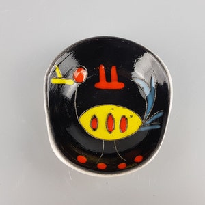 May include: A small black ceramic dish with a stylized bird design. The bird is yellow with red accents and blue feathers. The dish is perfect for serving small snacks or condiments.