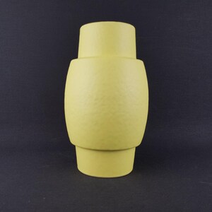 May include: A pale yellow ceramic vase with a textured surface. The vase has a cylindrical shape with a wider, rounded middle section and a narrower top and base. The background is a dark gray.