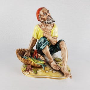 May include: A ceramic figurine of a fisherman sitting with a basket of fish. He is wearing a red hat, a yellow shirt, and green pants. The figurine is painted in realistic detail and has a rustic charm.