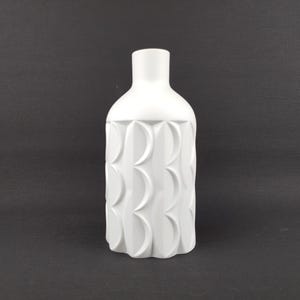 May include: A white ceramic vase with a textured, repeating pattern of curved shapes on the body. The vase has a narrow neck and a rounded shoulder. The background is a solid black.