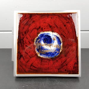 May include: Square ceramic tile with a vibrant red background and a central design featuring a blue and white floral pattern. The tile is framed in a white border, creating a striking contrast.