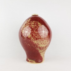 Peut inclure: Un vase en céramique bulbeux avec une finition brillante, présentant un vernis rouge profond superposé sur une base beige mouchetée. Le vase a un col étroit et un corps large et arrondi qui se rétrécit vers une base plate, avec un motif tacheté.