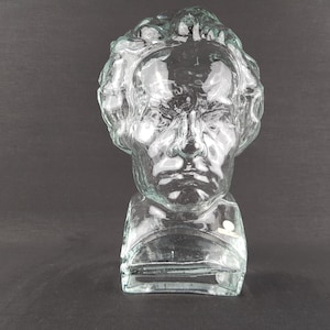 May include: A clear glass bust sculpture depicting a person's head and shoulders. The sculpture has detailed facial features and curly hair. The bust is set against a dark background.