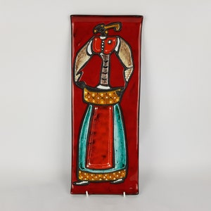 May include: A red ceramic wall plaque with a stylized figure of a person wearing a red, yellow, and blue outfit. The figure is outlined in black and has a white background.