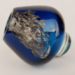 Puede incluir: Un tap&oacute;n de botella de vidrio azul con un dise&ntilde;o de remolino plateado. El tap&oacute;n tiene una base de vidrio transparente.