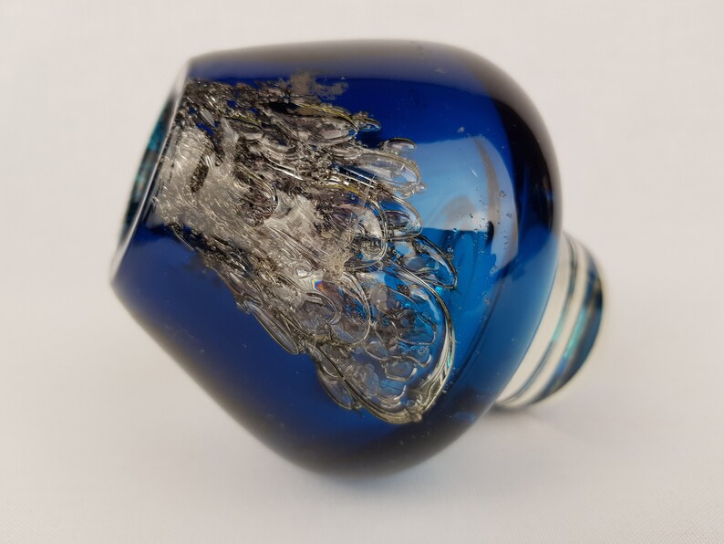 Puede incluir: Un tap&oacute;n de botella de vidrio azul con un dise&ntilde;o de remolino plateado. El tap&oacute;n tiene una base de vidrio transparente.