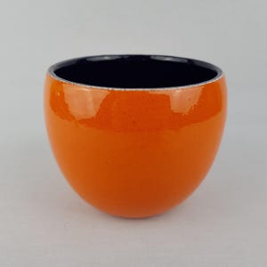 Puede incluir: Un pequeño cuenco de cerámica redondeado con un acabado exterior brillante de color naranja vibrante y un interior de color negro profundo que contrasta. El borde del cuenco es fino y liso, mostrando una abertura circular limpia.